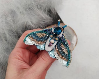 May include: A blue and white beaded moth brooch with iridescent sequins and a large blue gemstone in the center.