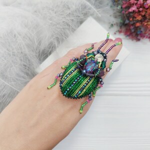 Peut inclure: Une broche en forme de scarab&eacute;e en perles vertes et violettes avec une grosse pierre pr&eacute;cieuse iris&eacute;e au centre. La broche est sur la main d'une personne.