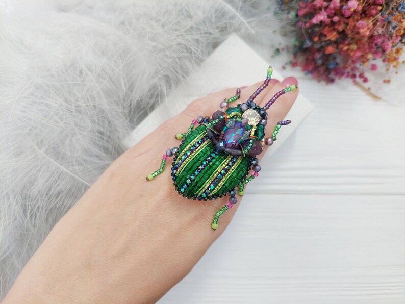 Peut inclure: Une broche en forme de scarab&eacute;e en perles vertes et violettes avec une grosse pierre pr&eacute;cieuse iris&eacute;e au centre. La broche est sur la main d'une personne.