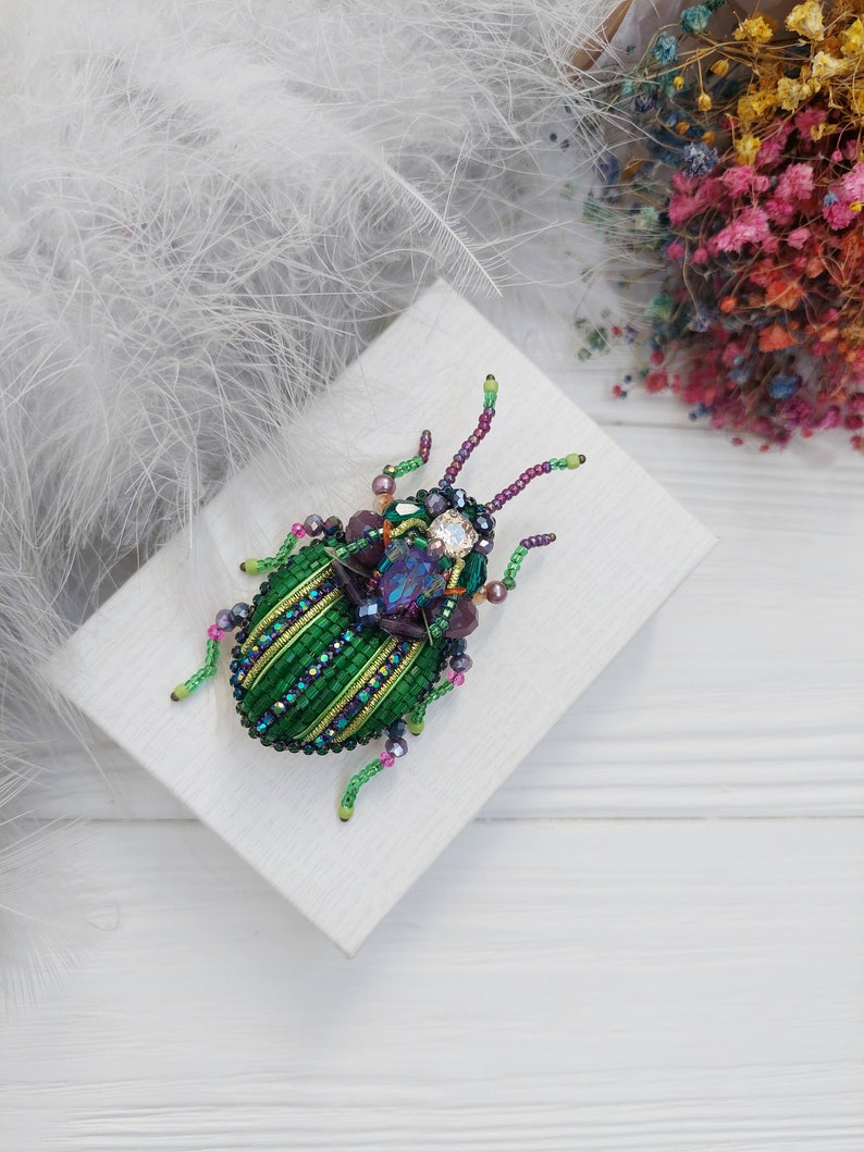 Peut inclure: Une broche en forme de scarab&eacute;e en perles vertes et violettes avec un strass transparent au centre. La broche est sur une bo&icirc;te blanche.