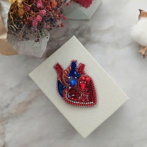 May include: A red and blue beaded heart brooch with a large blue rhinestone in the center. The brooch is displayed on a white box.