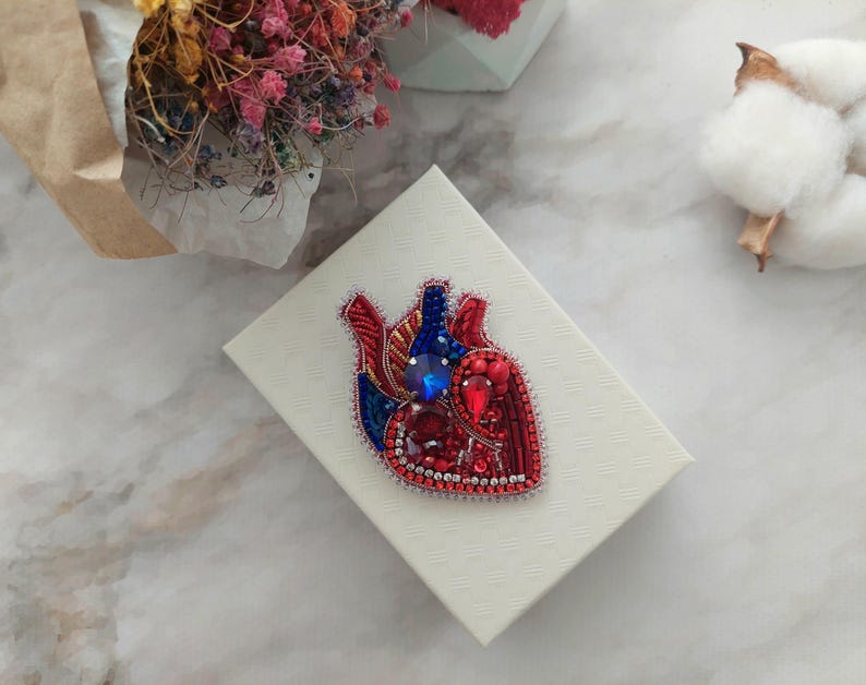 May include: A red and blue beaded heart brooch with a large blue rhinestone in the center. The brooch is displayed on a white box.