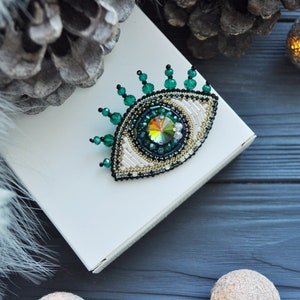 May include: A green and white beaded eye brooch with a large iridescent crystal in the center. The brooch is on a white box and a dark wood background.