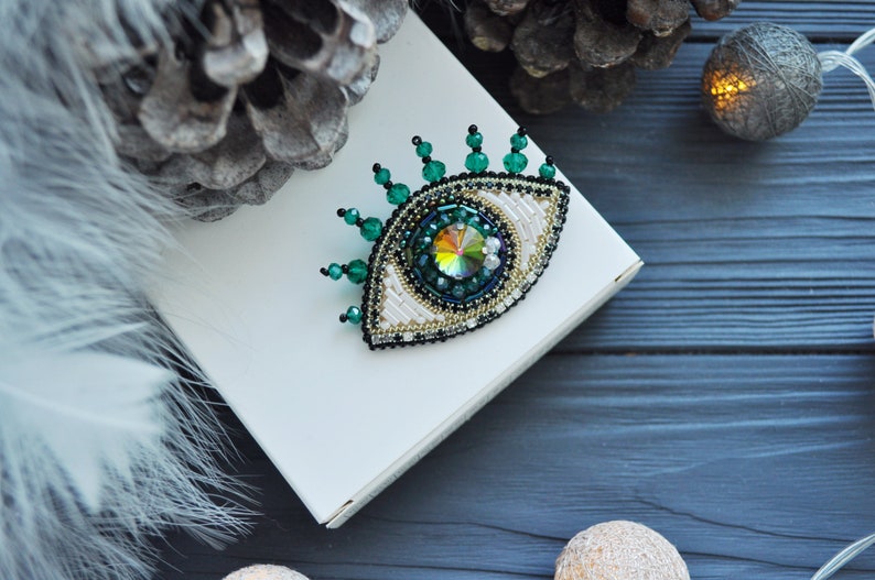 May include: A green and white beaded eye brooch with a large iridescent crystal in the center. The brooch is on a white box and a dark wood background.