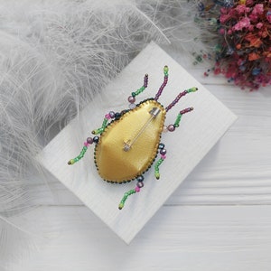 Peut inclure: Une broche en forme d'insecte en perles dor&eacute;es et vertes avec une &eacute;pingle au dos. La broche est sur une surface blanche.