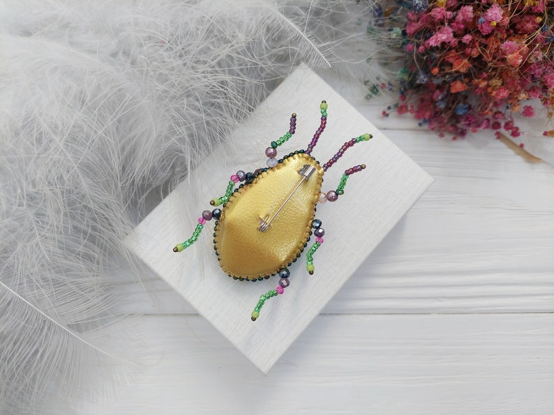 Peut inclure: Une broche en forme d'insecte en perles dor&eacute;es et vertes avec une &eacute;pingle au dos. La broche est sur une surface blanche.