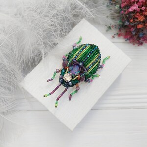 Peut inclure: Une broche en forme de scarab&eacute;e en perles vertes et violettes avec un strass transparent au centre. La broche est sur une surface blanche.