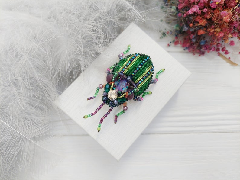 Peut inclure: Une broche en forme de scarab&eacute;e en perles vertes et violettes avec un strass transparent au centre. La broche est sur une surface blanche.