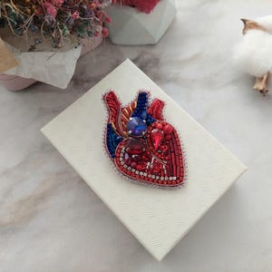May include: A red and blue beaded heart brooch with rhinestones. The brooch is on a white box.