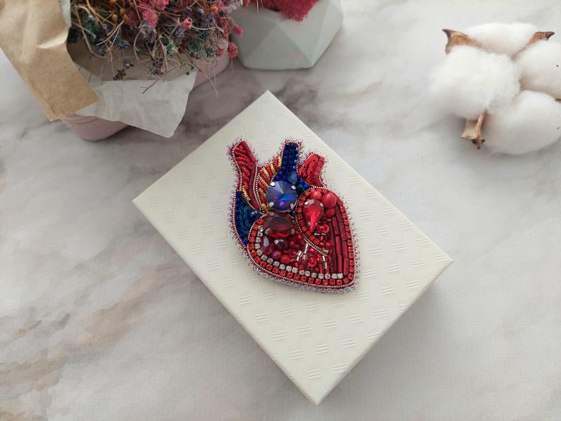 May include: A red and blue beaded heart brooch with rhinestones. The brooch is on a white box.