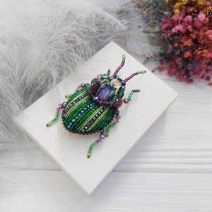 Peut inclure: Une broche en forme de scarab&eacute;e en perles vertes et violettes avec des accents iris&eacute;s. La broche est sur une surface blanche.