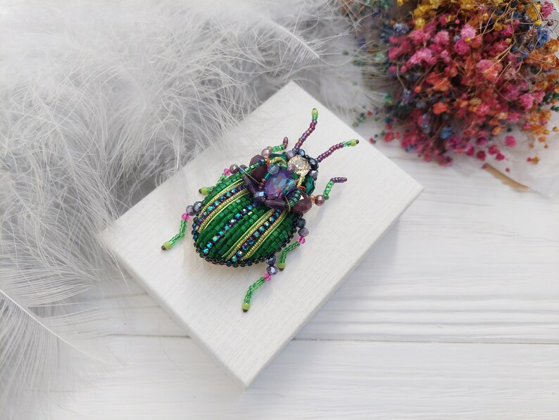 Peut inclure: Une broche en forme de scarab&eacute;e en perles vertes et violettes avec des accents iris&eacute;s. La broche est sur une surface blanche.