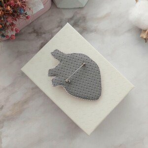 May include: A gray heart-shaped brooch with a pin on the back. The brooch is on a white box with a textured surface.