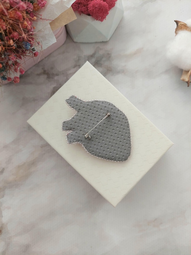 May include: A gray heart-shaped brooch with a pin on the back. The brooch is on a white box with a textured surface.