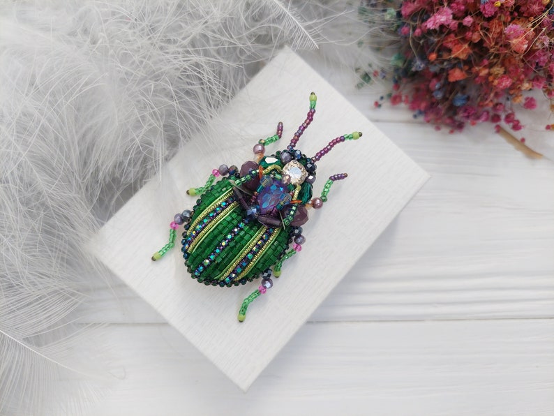Peut inclure: Une broche en forme de scarab&eacute;e en perles vertes et violettes avec un grand bijou iridescent au centre. La broche est sur un fond blanc.
