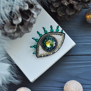 May include: A green and white beaded eye brooch with a sparkly iridescent center. The brooch is on a white box and a dark blue wooden surface.