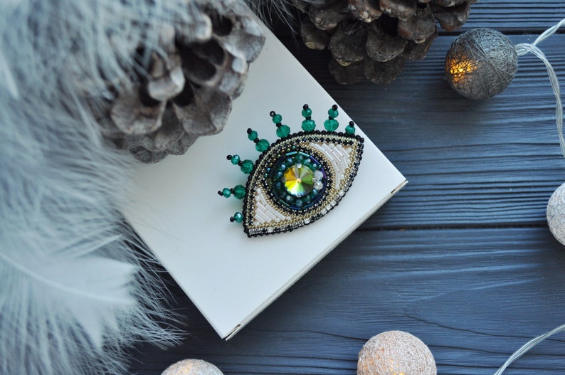 May include: A green and white beaded eye brooch with a sparkly iridescent center. The brooch is on a white box and a dark blue wooden surface.