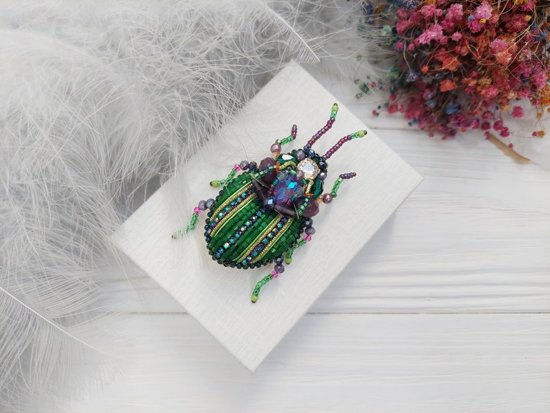 Peut inclure: Une broche en forme de scarab&eacute;e en perles vertes et violettes avec un corps orn&eacute; de bijoux. La broche est sur une surface blanche.