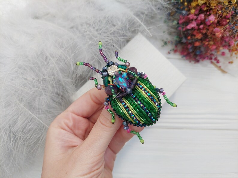 Peut inclure: Une broche en forme de scarab&eacute;e en perles vertes et violettes avec un strass transparent sur le dos. Le scarab&eacute;e a un motif ray&eacute; et est fait de petites perles.