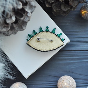 May include: A green and gold eye-shaped brooch with a pin on the back. The brooch is on a white surface with a pine cone and string lights in the background.