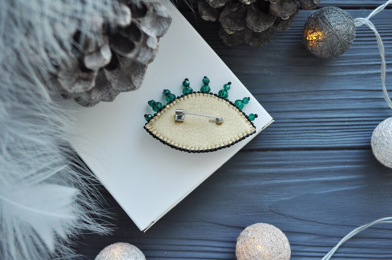May include: A green and gold eye-shaped brooch with a pin on the back. The brooch is on a white surface with a pine cone and string lights in the background.