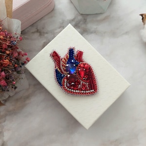 May include: A red, blue, and gold beaded heart brooch with rhinestones. The brooch is on a white box with a textured surface.