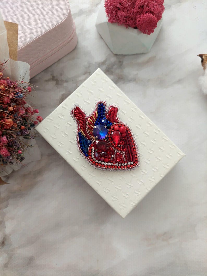May include: A red, blue, and gold beaded heart brooch with rhinestones. The brooch is on a white box with a textured surface.