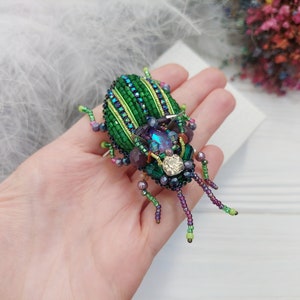 Peut inclure: Une broche en forme de scarab&eacute;e en perles vertes et violettes avec des strass iris&eacute;s. Le scarab&eacute;e a un design d&eacute;taill&eacute; avec des rayures et un corps textur&eacute;.