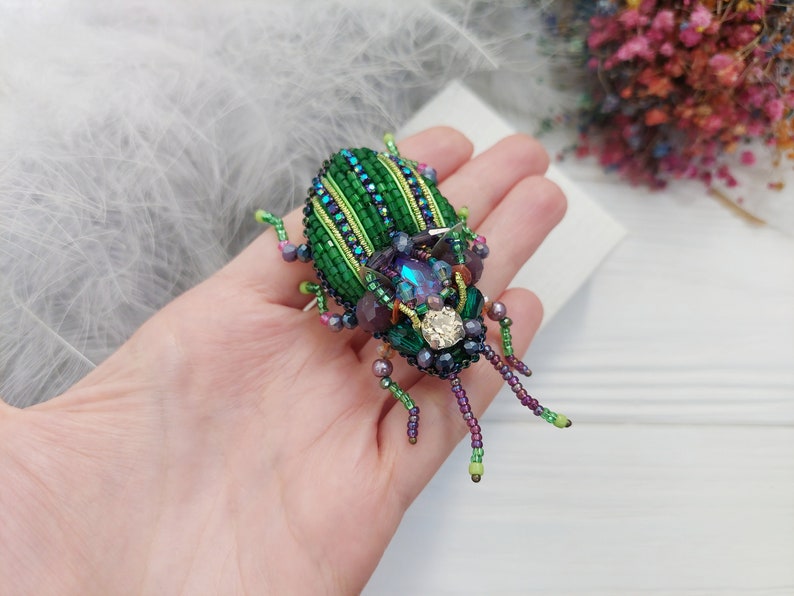 Peut inclure: Une broche en forme de scarab&eacute;e en perles vertes et violettes avec des strass iris&eacute;s. Le scarab&eacute;e a un design d&eacute;taill&eacute; avec des rayures et un corps textur&eacute;.