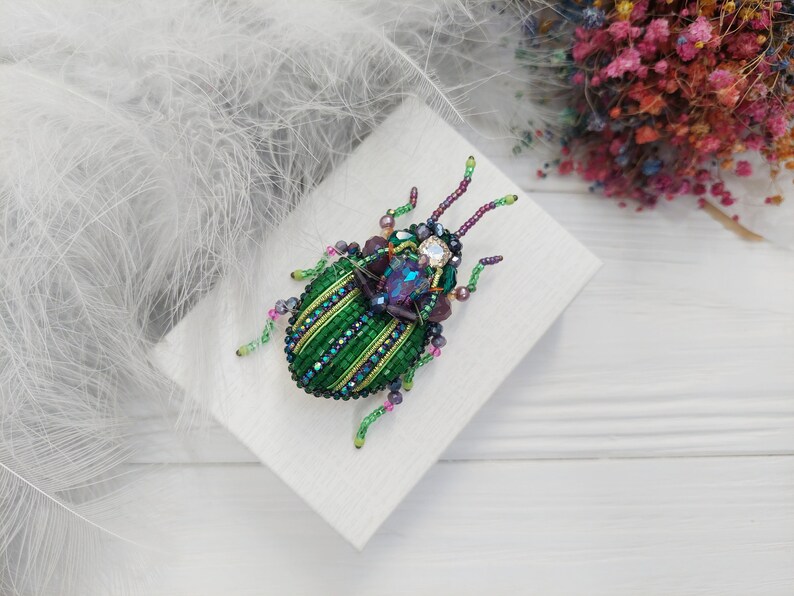 Peut inclure: Une broche en forme de scarab&eacute;e en perles vertes et violettes avec des accents iris&eacute;s. La broche est sur un fond blanc.