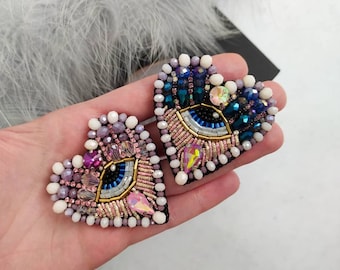 May include: Two heart-shaped beaded brooches featuring an eye design. The brooches are crafted with pink, white, blue, and gold beads.