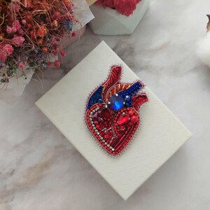 May include: A red, blue, and silver beaded heart brooch with a blue rhinestone in the center. The brooch is on a white background.