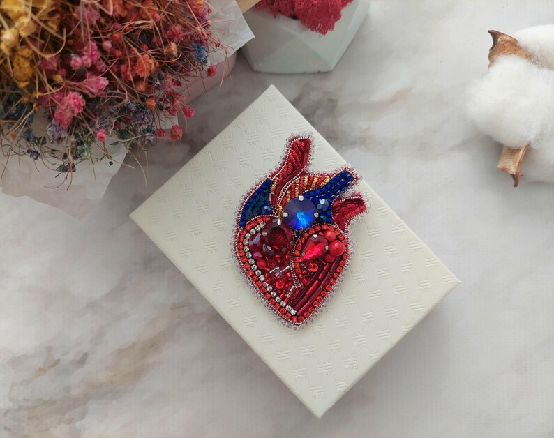 May include: A red, blue, and silver beaded heart brooch with a blue rhinestone in the center. The brooch is on a white background.