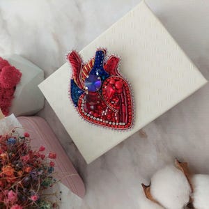 May include: A red, blue, and silver beaded heart-shaped brooch. The heart is detailed with a blue gem in the center and red and silver beads.