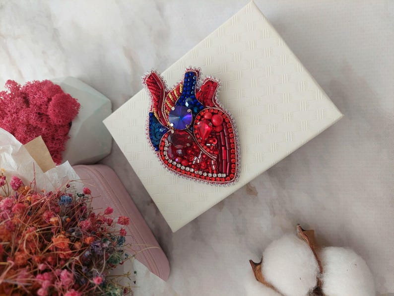 May include: A red, blue, and silver beaded heart-shaped brooch. The heart is detailed with a blue gem in the center and red and silver beads.