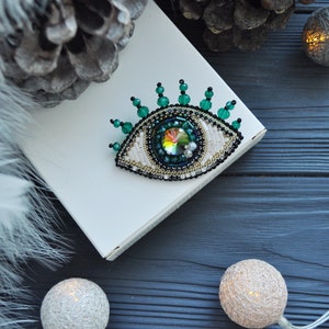 May include: A beaded eye brooch with a green and white design. The eye is surrounded by green beads and has a large iridescent crystal in the center. The brooch is on a white surface.