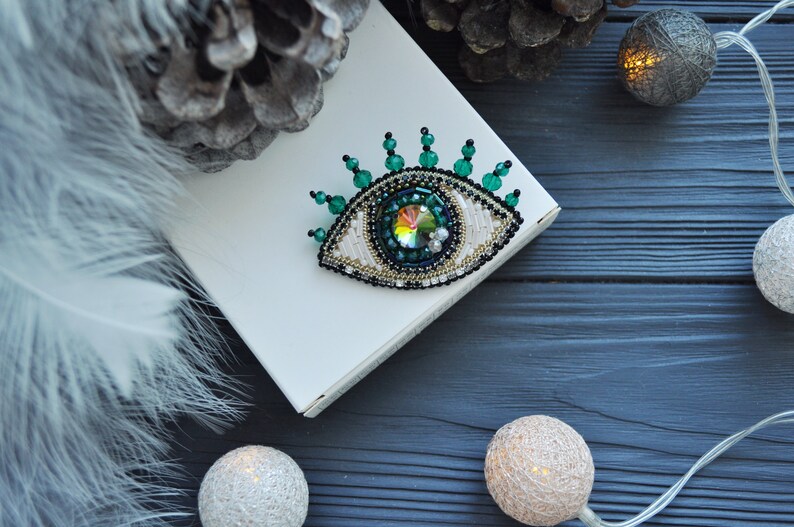 May include: A beaded eye brooch with a green and white design. The eye is surrounded by green beads and has a large iridescent crystal in the center. The brooch is on a white surface.