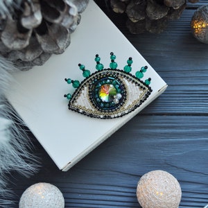 May include: A green and white beaded eye brooch with a crystal center. The brooch is on a white surface with a pine cone and string lights in the background.