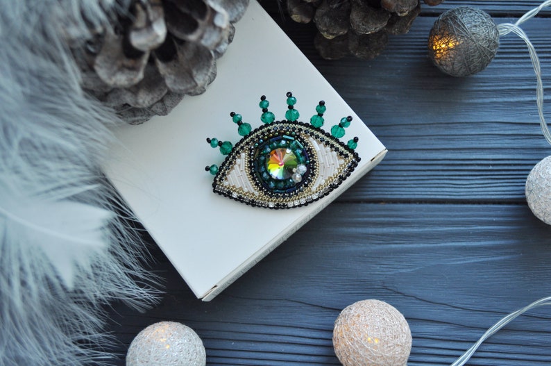 May include: A green and white beaded eye brooch with a crystal center. The brooch is on a white surface with a pine cone and string lights in the background.