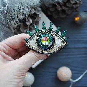 May include: A beaded eye brooch with a green and white design. The eye is surrounded by green beads and has a iridescent jewel in the center.