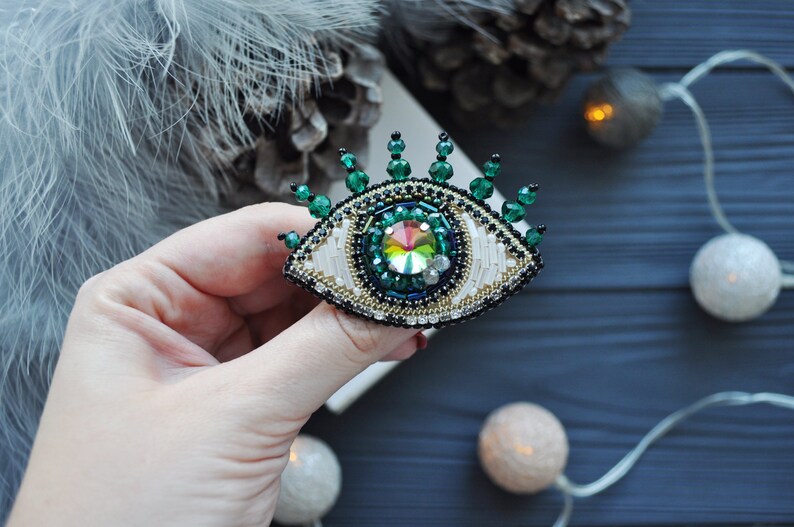 May include: A beaded eye brooch with a green and white design. The eye is surrounded by green beads and has a iridescent jewel in the center.