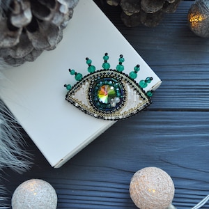 May include: A green and gold beaded eye brooch with a crystal center. The brooch is on a white box with a fuzzy white background.