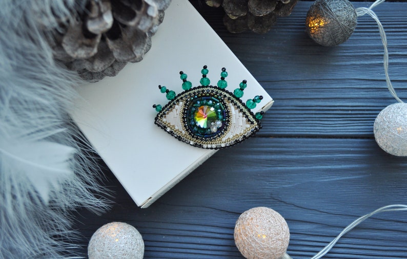 May include: A green and gold beaded eye brooch with a crystal center. The brooch is on a white box with a fuzzy white background.
