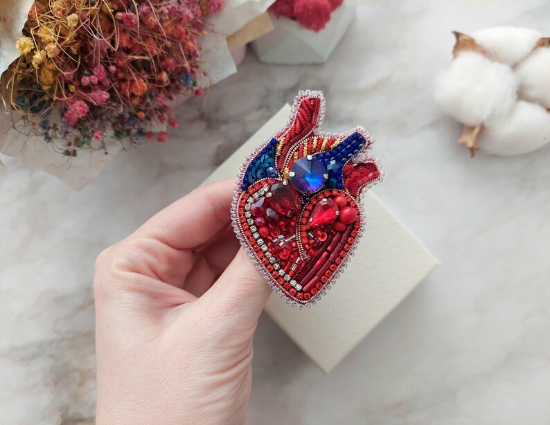 May include: A red, blue, and gold beaded heart brooch with a detailed anatomical design. The heart is embellished with rhinestones and red beads.
