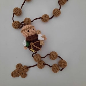 May include: A crocheted rosary with brown beads and a brown cross. The rosary features a crocheted figure of Saint Anthony holding a baby.
