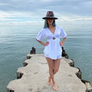 Pode incluir: Uma mulher vestindo um vestido branco de croch&ecirc; e um chap&eacute;u de abas largas com bordados coloridos est&aacute; em p&eacute; em um p&iacute;er de concreto com vista para um corpo d'&aacute;gua.
