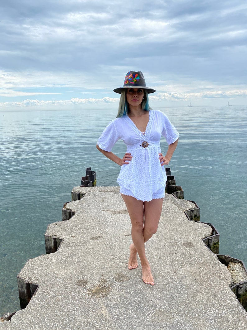 Pode incluir: Uma mulher vestindo um vestido branco de croch&ecirc; e um chap&eacute;u de abas largas com bordados coloridos est&aacute; em p&eacute; em um p&iacute;er de concreto com vista para um corpo d'&aacute;gua.