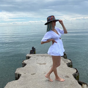 Pode incluir: Uma mulher vestindo um vestido branco e um chap&eacute;u de abas largas colorido est&aacute; em p&eacute; em um p&iacute;er de concreto com vista para um corpo de &aacute;gua. O c&eacute;u est&aacute; nublado e a &aacute;gua est&aacute; calma.