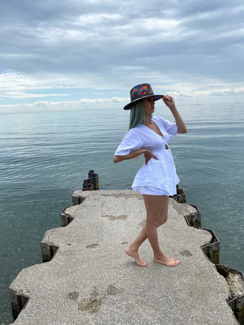 Pode incluir: Uma mulher vestindo um vestido branco e um chap&eacute;u de abas largas colorido est&aacute; em p&eacute; em um p&iacute;er de concreto com vista para um corpo de &aacute;gua. O c&eacute;u est&aacute; nublado e a &aacute;gua est&aacute; calma.