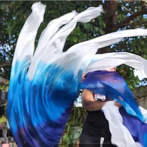 May include: A flowing fabric display in white and blue hues, creating a dynamic, wave-like effect. The fabric appears lightweight and airy, with a colour gradient from white to deep blue. The image is set against a blurred background of green foliage.